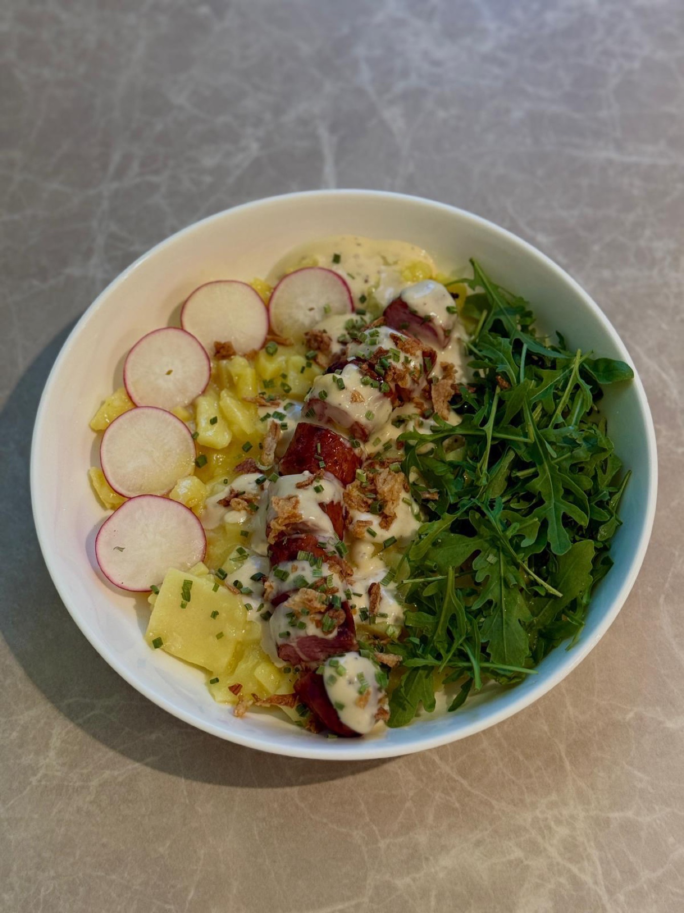 Frisch angerichtete Bowl mit Kartoffeln, Wurst, Rucola und Dressing.